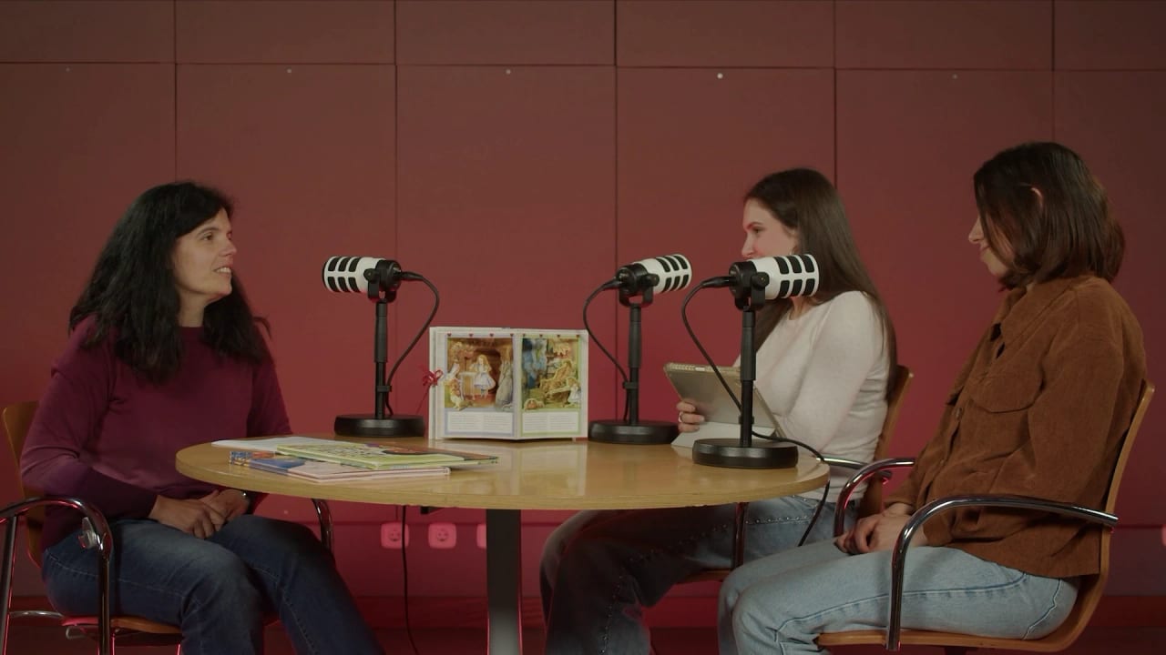 Conversa no estúdio do Hora do Saber, com a convidada e as apresentadoras à mesa, entre microfones e um livro pop-up